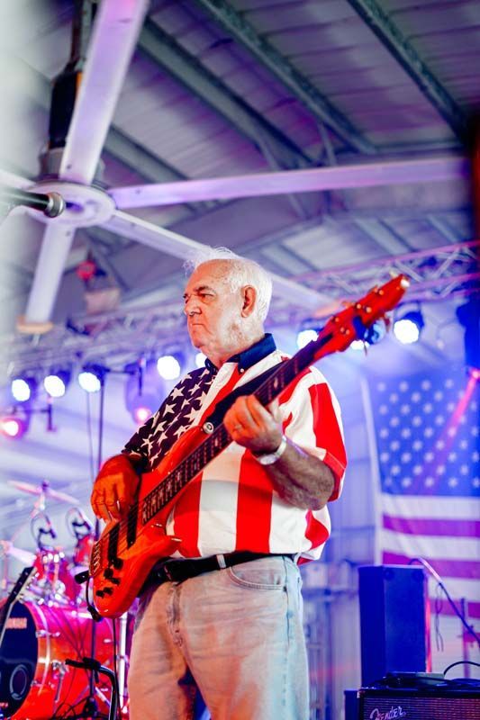 Musician playing a red electric guitar onstage under purple lights, with a large American flag backdrop.