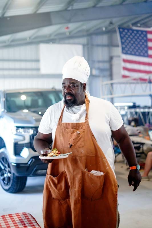 Chef in a white hat and orange apron carries a plate inside a garage with a truck and U.S. flag.