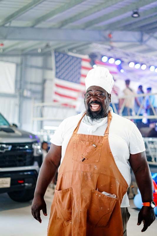 Smiling man in orange overalls and white hat standing in a garage with a car and American flag behind him