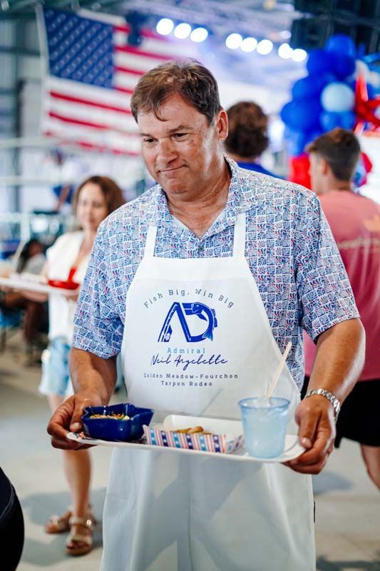 Man in a floral shirt and apron serving food on a tray at a festive indoor event with American flags and balloons.