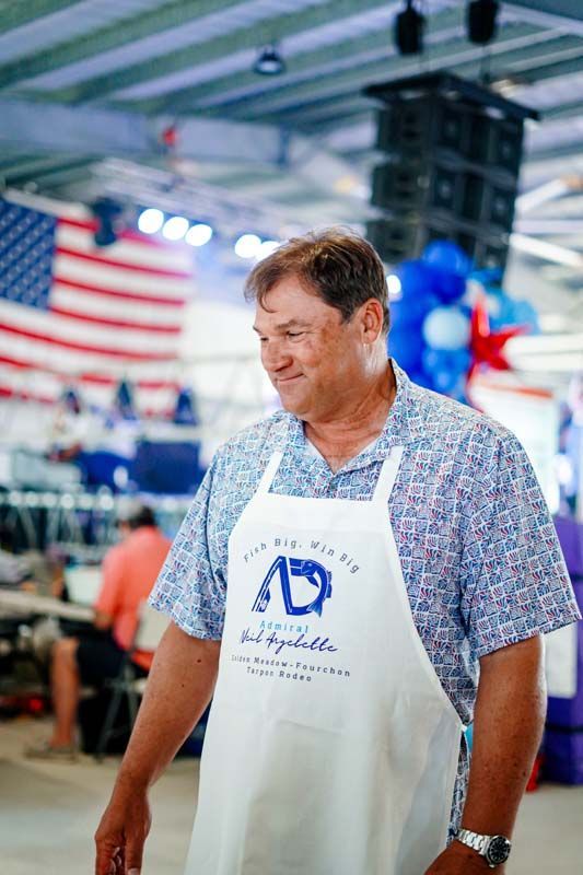 Man smiling in a white apron at an indoor event with a large American flag in the background