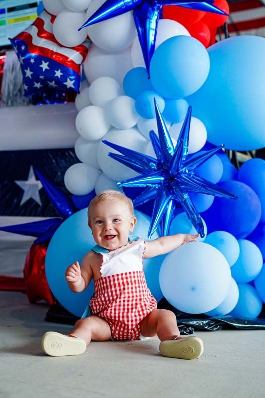 Smiling baby in red checkered outfit sitting by blue and white balloons and star decorations