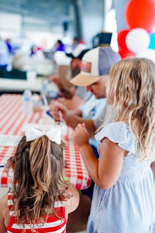 Children seated at a red-and-white checkered table, eating at a festive indoor party with balloons.