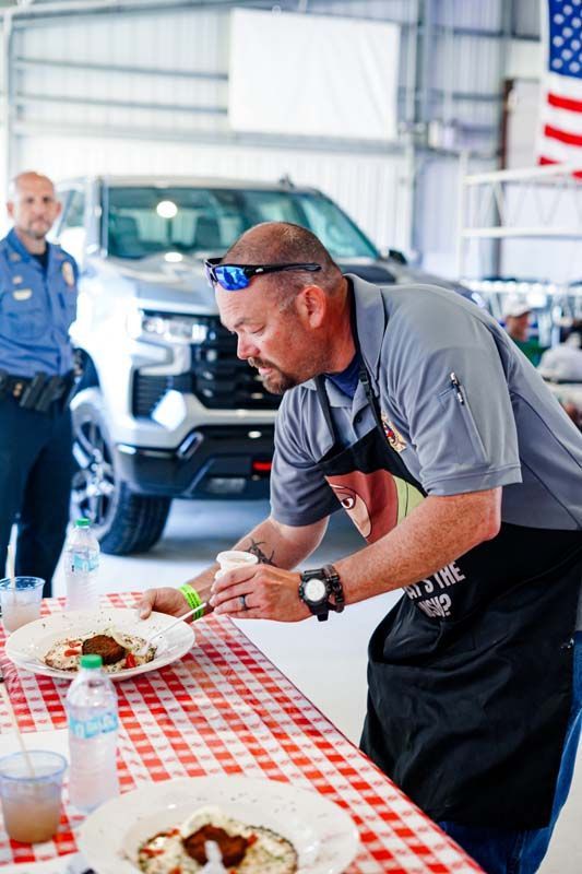 Man serving food at a table in a hangar, with a truck and American flag in the background
