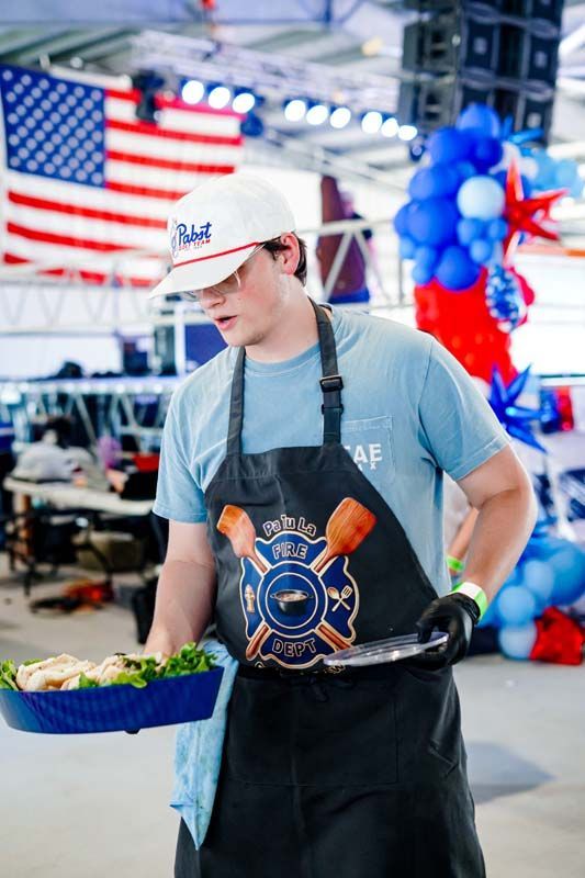 Person in apron and gloves carrying a tray of greens at a festive indoor market with balloons and an American flag.