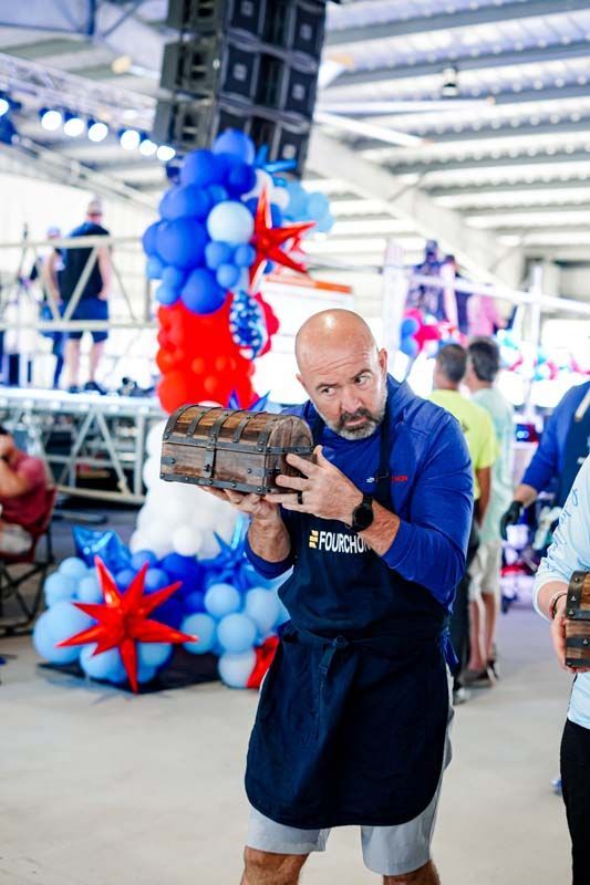 Man holding a wooden sculpture indoors beside a red, white, and blue balloon display