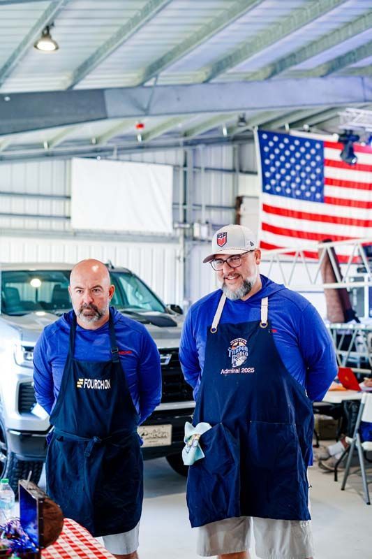 Two men in blue aprons standing in a garage with a truck and a large American flag in the background