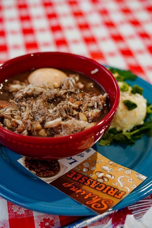 Red bowl of hearty stew on a blue plate with crackers and a checkered tablecloth.