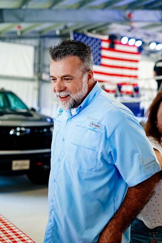 Man in a light blue shirt standing in a showroom with a black car and an American flag in the background