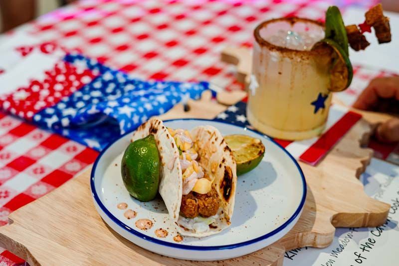 Pecan pie slice with whipped cream and lime on a plate, beside a margarita on a red checkered picnic table.