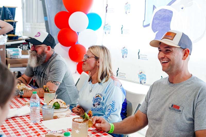 Three people seated at a table with food and drinks at a festive indoor event, smiling and talking