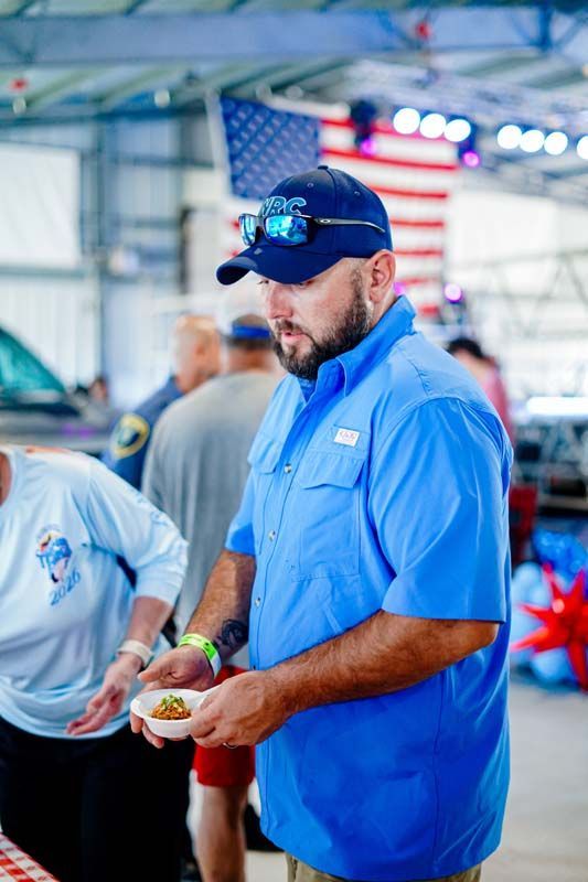 Man in a blue shirt and cap holding a plate of food at an indoor event with an American flag in the background