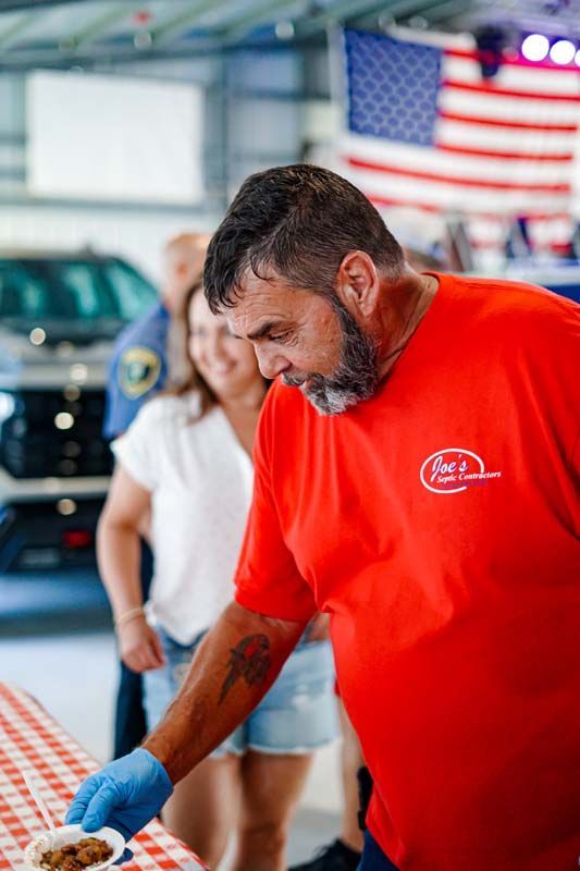 Man in a red shirt and blue gloves works at a table in a workshop with a car and U.S. flag in the background