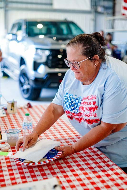 Woman making something at a red-and-white checkered table in a workshop, with a truck in the background