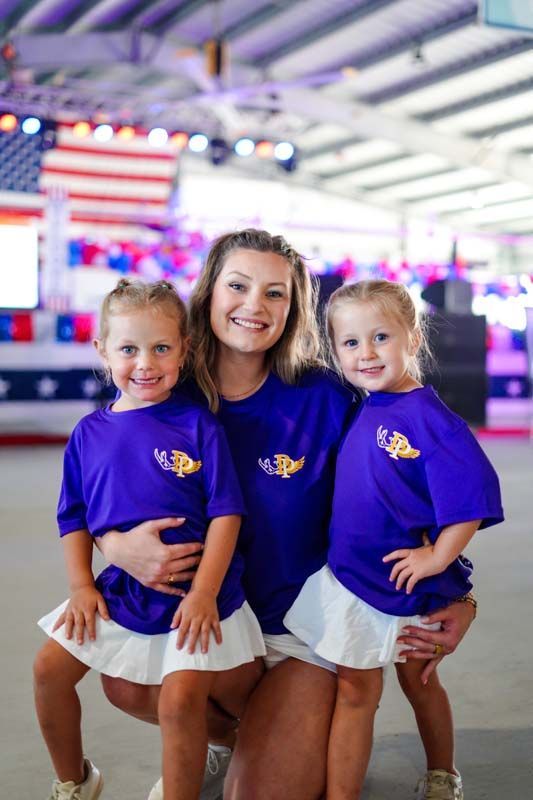 Three smiling children in purple shirts and white skirts posing indoors on a rink-like floor