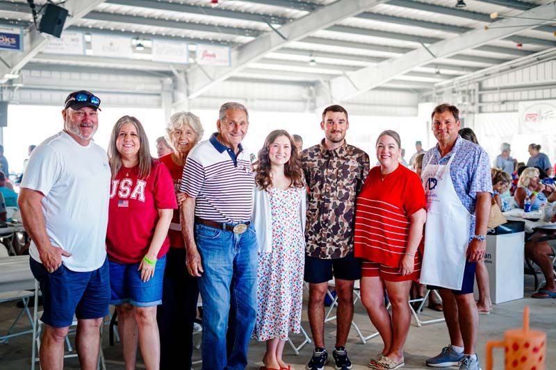Group of people posing in a bright indoor venue with exposed beams and picnic tables, smiling at the camera.