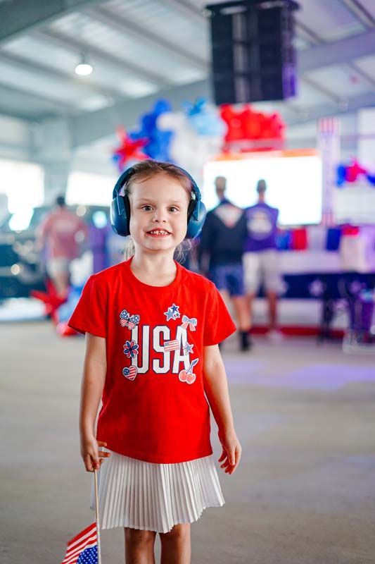 Child in red shirt and striped skirt standing indoors, wearing headphones with a blurred event crowd behind.