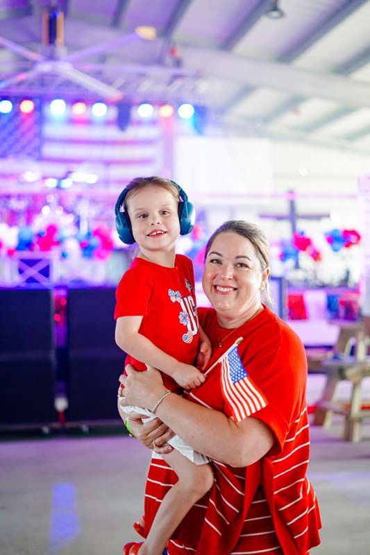 Woman holding a child indoors, both in red shirts, with an American flag in the background