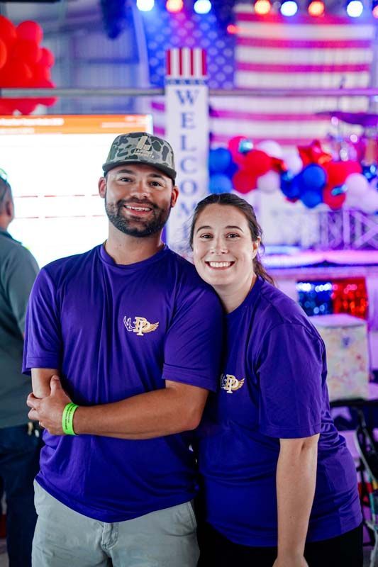 Two people in purple shirts smile at a festive booth with balloons and an American flag backdrop.