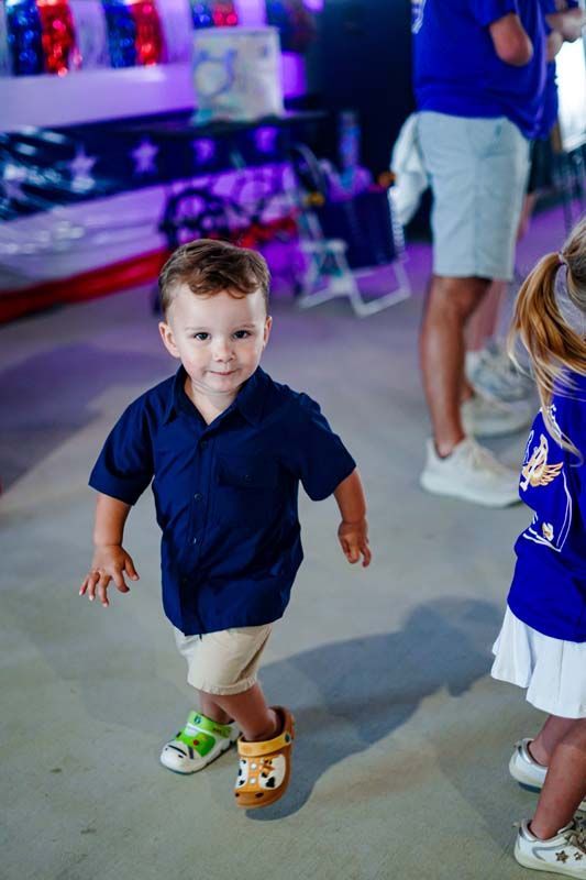Toddler in a navy shirt and tan shorts walking indoors at a party, with colorful lights in the background