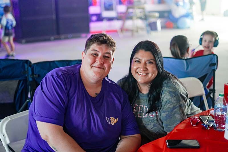 Two people smiling at a table, wearing purple and camo shirts, with red tablecloth and drinks.