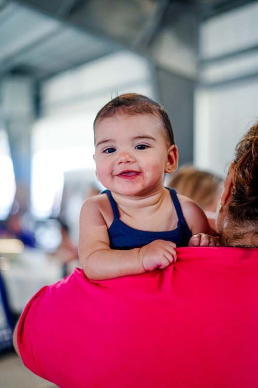 Smiling baby in a navy swimsuit being held over a bright pink shoulder in a blurred indoor setting