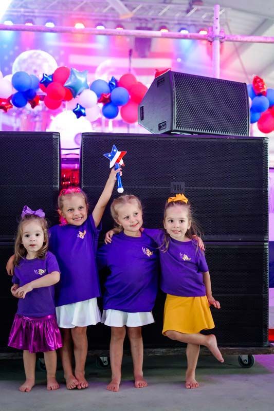 Four children in purple shirts pose on stage with balloons and speakers behind them
