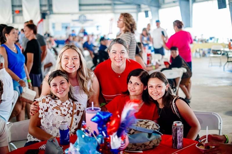 Group smiling at a party table with red tablecloth, balloons, and snacks in a bright indoor hall