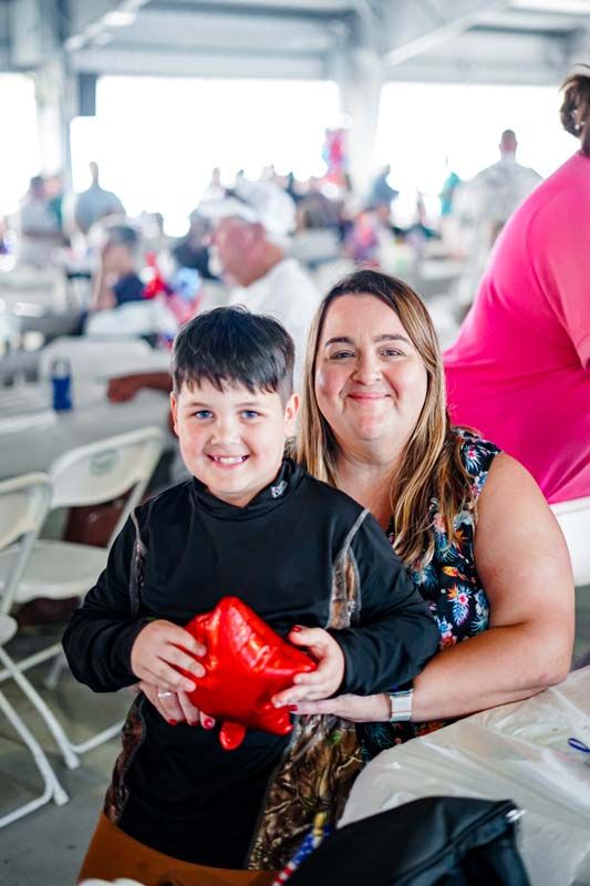 Smiling child holding a red heart balloon while seated beside a woman at a bright indoor event