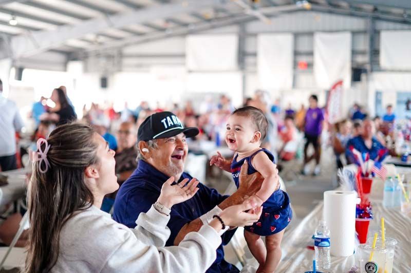 Woman lifts smiling child at a crowded indoor event, with people seated at tables in the background.