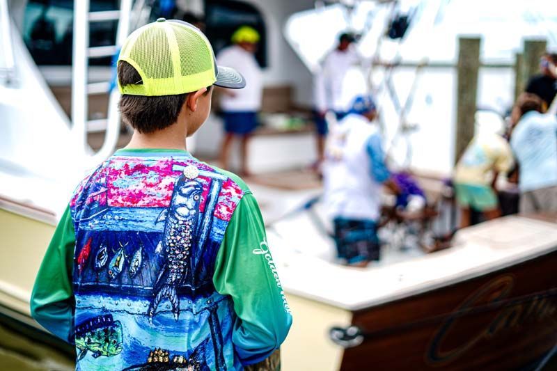 A young boy wearing a green shirt and a yellow hat is standing in front of a boat.