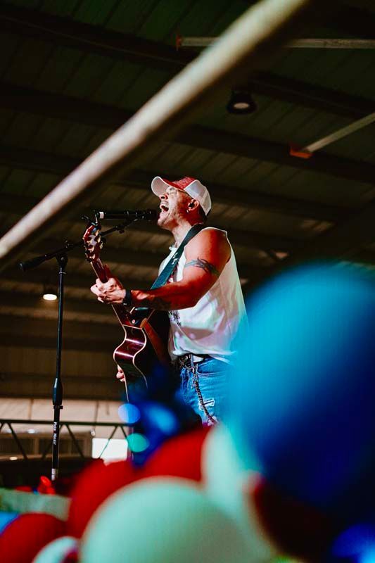 Musician playing guitar onstage under warm lights, with blurred balloons in the foreground