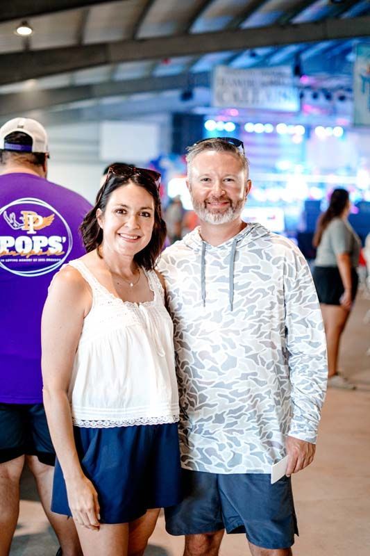 Two people smiling indoors at a busy event venue, standing side by side in casual summer clothes.