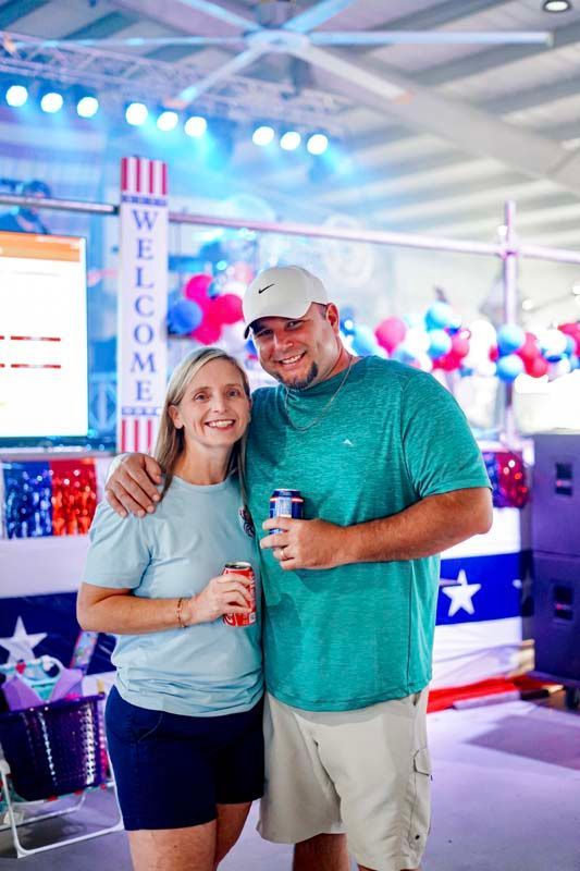 Smiling couple holding drinks at a patriotic indoor event with blue lights and a “WELCOME” sign.
