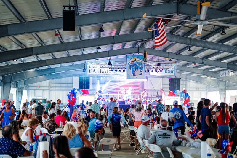 Crowded indoor rally with a stage, banners, blue-red balloons, and an American flag hanging overhead