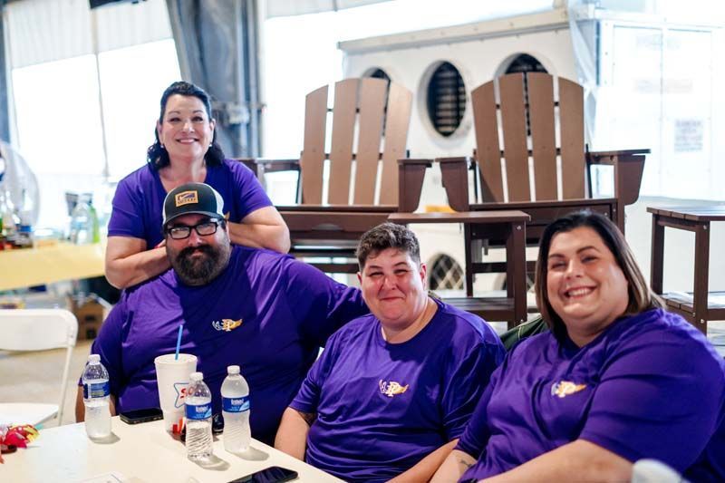 Four people in purple shirts smiling at a table indoors, with chairs stacked in the background.