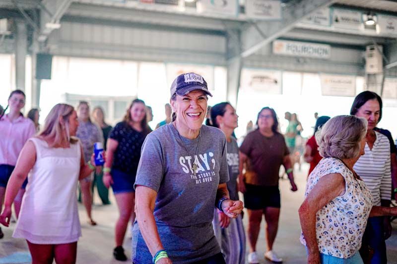 People dancing in a bright indoor hall, with one smiling woman in a gray shirt in the foreground