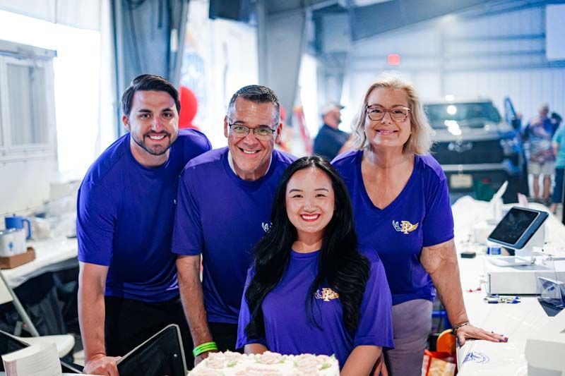 Four smiling people in purple shirts stand together in a bright office or lab setting.