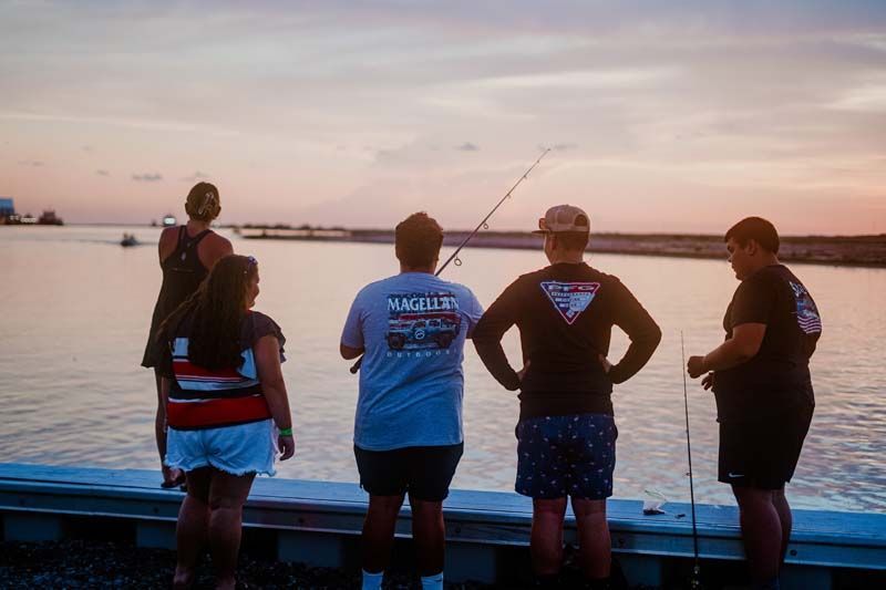 Five people stand on a dock at sunset, looking out over calm water with fishing rods.