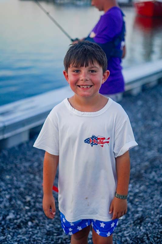 Smiling child in a white T-shirt and blue shorts standing by the water with a fishing rod in the background