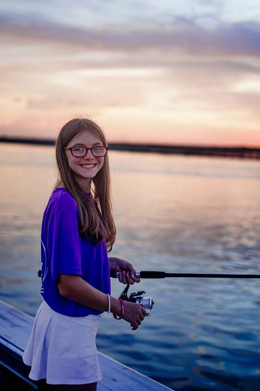 Person fishing by a calm lake at sunset, wearing a purple top and white skirt, smiling at the camera.