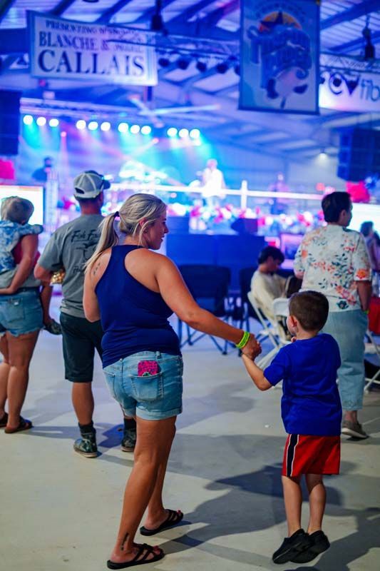 Crowd walking past bright blue fair lights at night, with a woman and child holding a toy in the foreground