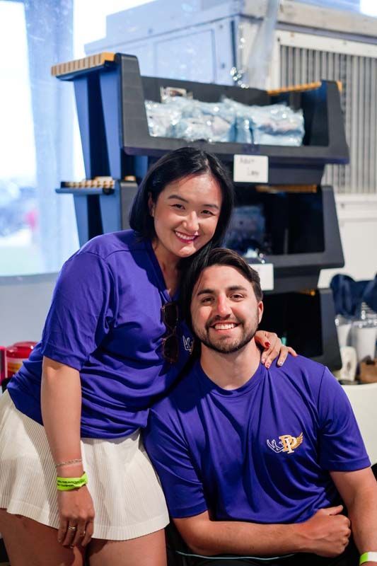 Two people in purple shirts smiling beside a black display stand in a bright indoor space