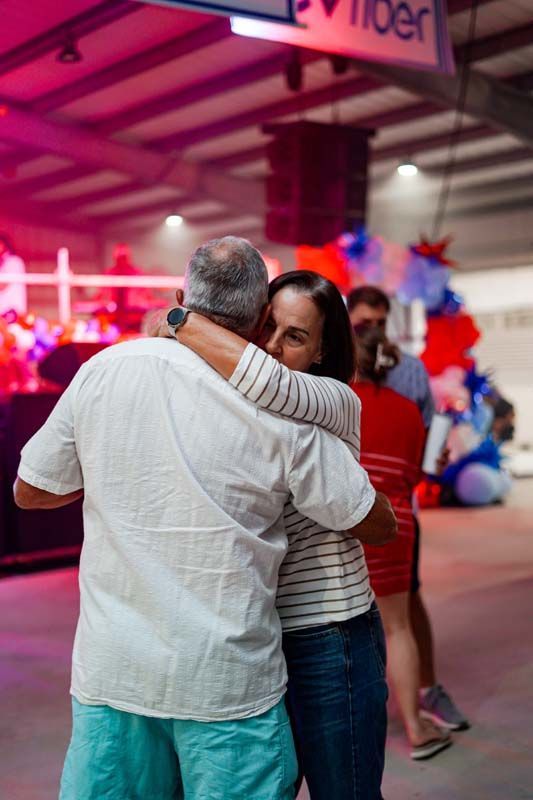Two people embracing at a brightly lit indoor event, with red and blue decorations in the background.