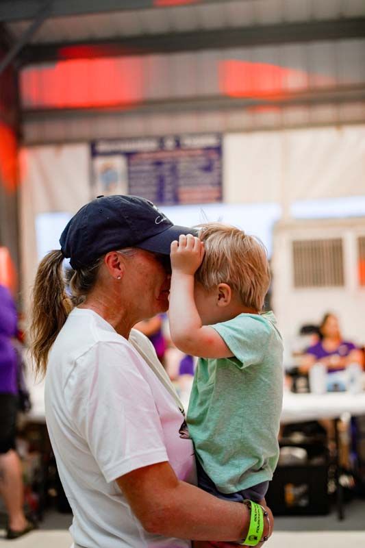 Person holding a child face-to-face indoors, both in casual clothes, with blurred people in the background