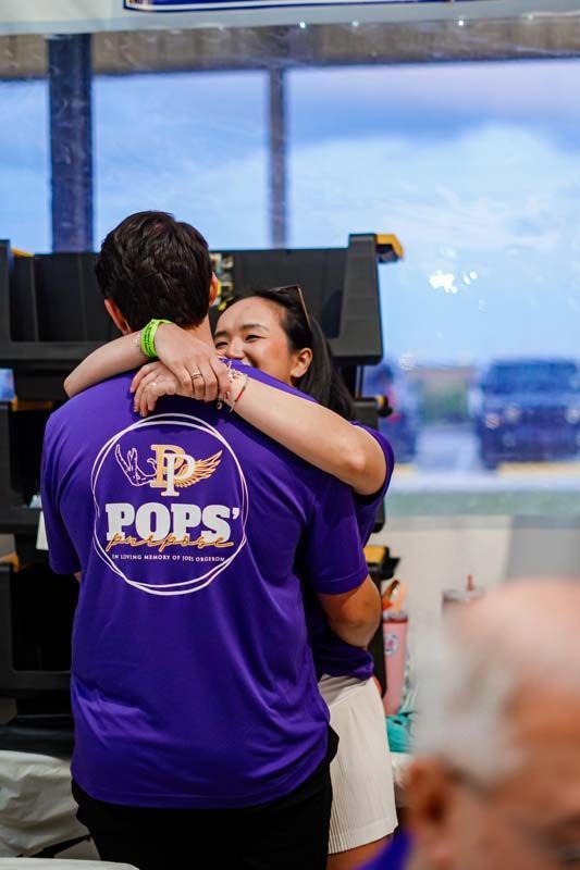 Two people hug indoors, one in a purple shirt with “POPS” on the back.