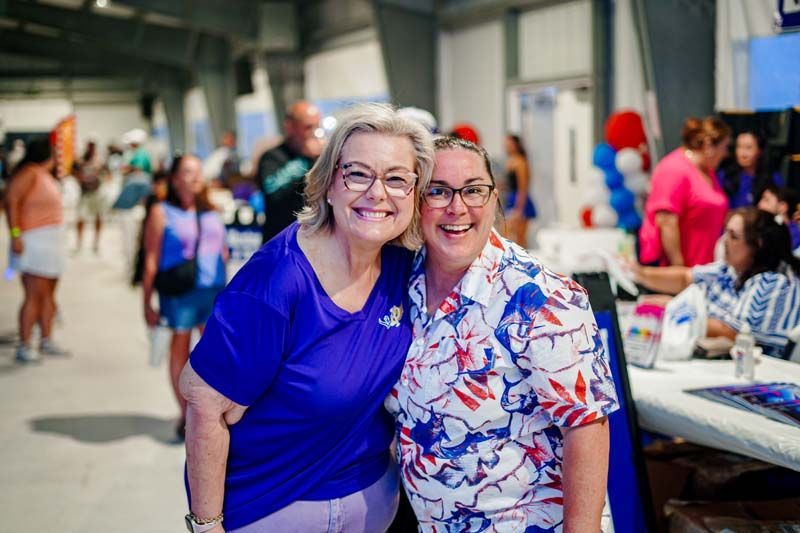 Two smiling people pose together at a busy indoor event, one in a blue shirt and one in a floral shirt.