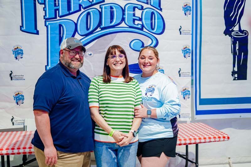Three people smiling in front of a “Thank You Rodeo” sign and checkered tablecloth