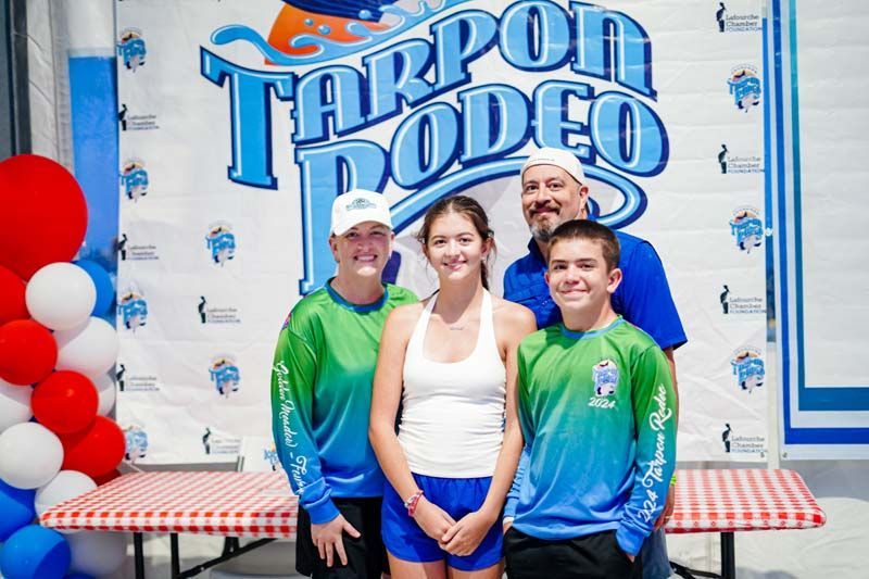 Three people posing in front of a Tarpon Rodeo banner with red and white balloons.