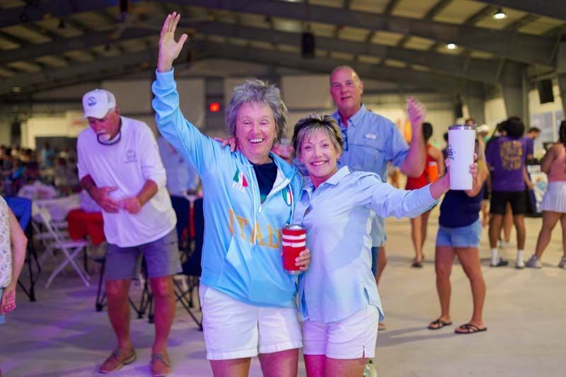 Two people in blue shirts smiling and waving at an indoor event, holding drinks.